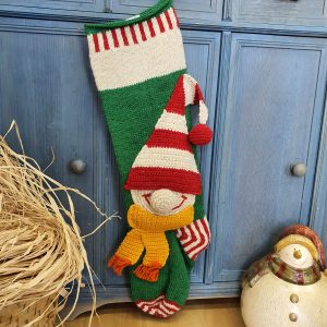Green and red hand-knit Christmas stocking with elf appliqué hanging on a blue cabinet
