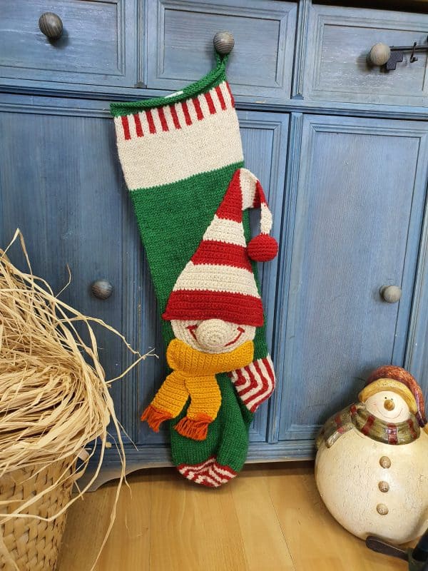 Green and red hand-knit Christmas stocking with elf appliqué hanging on a blue cabinet