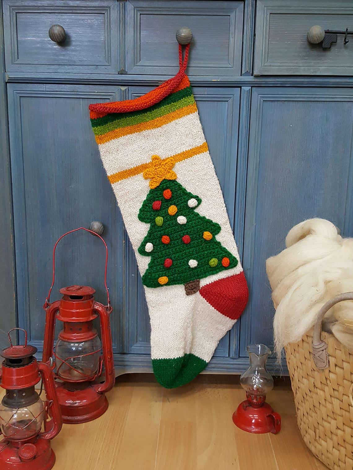 Hand-knitted Christmas stocking with green tree design, colorful ornaments, and striped cuff, hanging.
