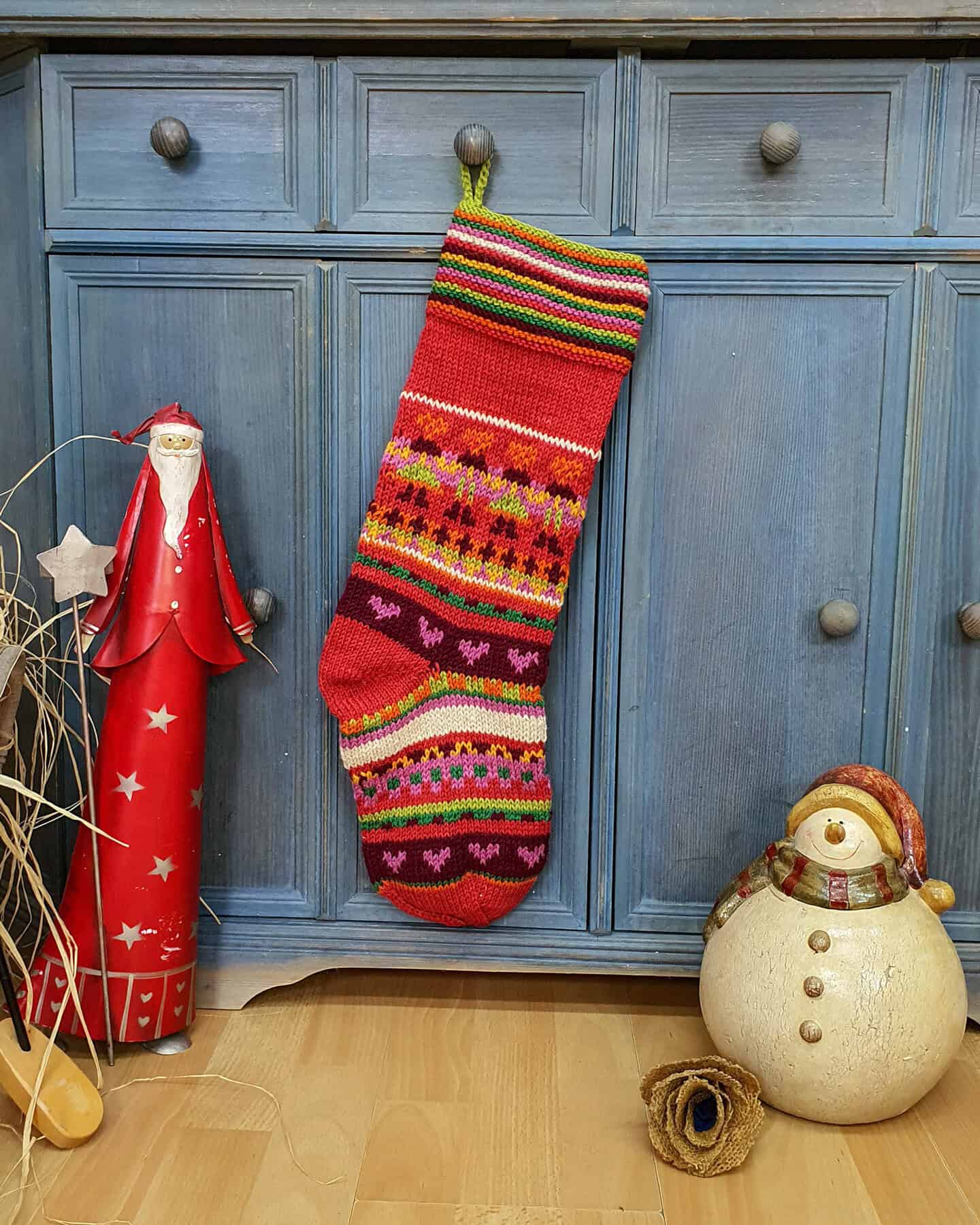 Hand-knitted Christmas stocking with colorful stripes and heart motifs, hung against a blue cabinet with festive decorations.