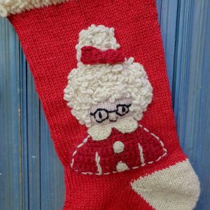 Close-up of hand-knitted Mrs. Claus Christmas stocking showing detailed face motif with glasses, curly hair, and festive hat.
