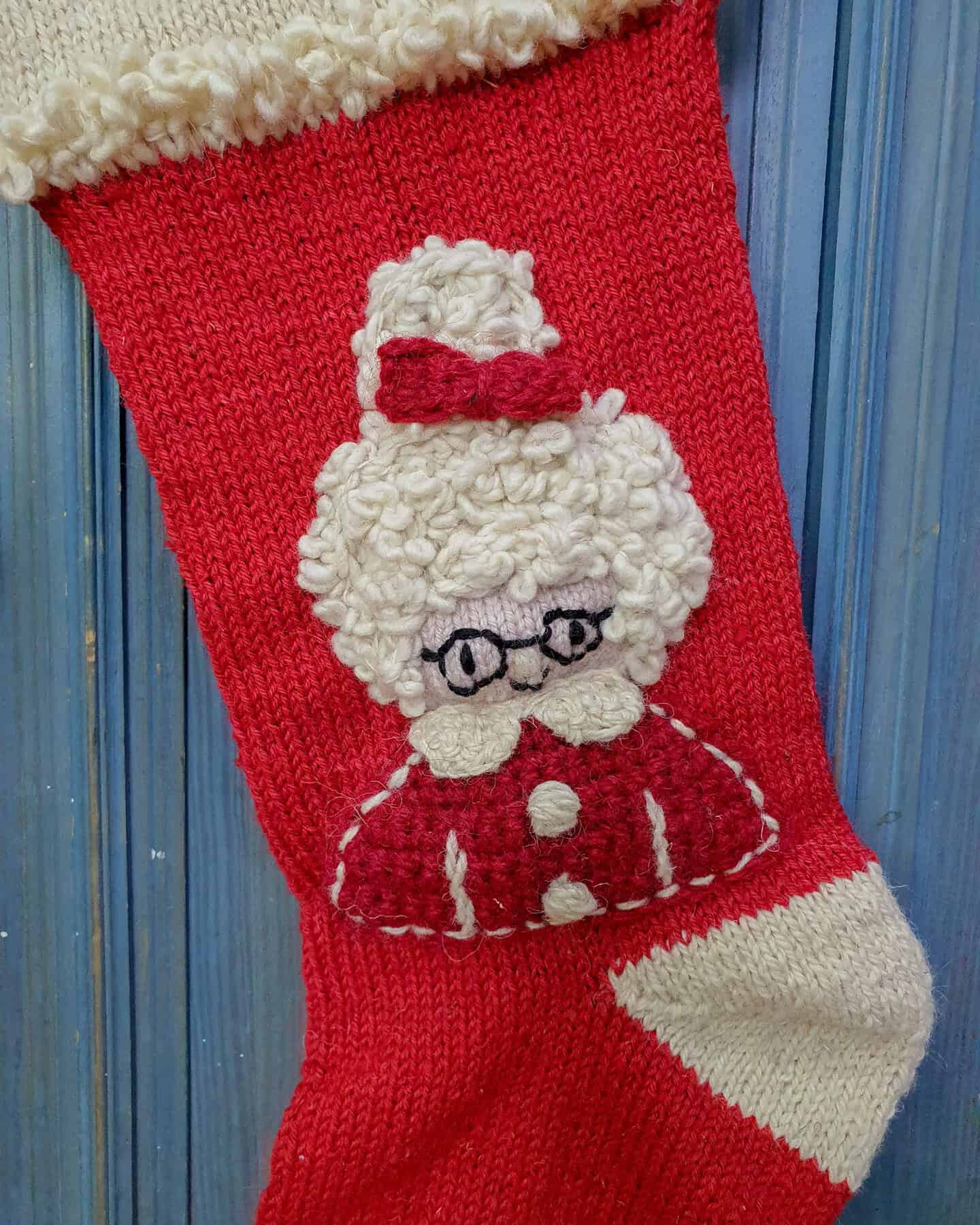 Close-up of hand-knitted Mrs. Claus Christmas stocking showing detailed face motif with glasses, curly hair, and festive hat.