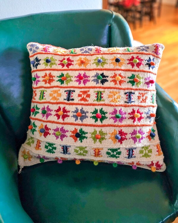 Hand-knitted pillow cover with colorful flower motifs and pom-poms