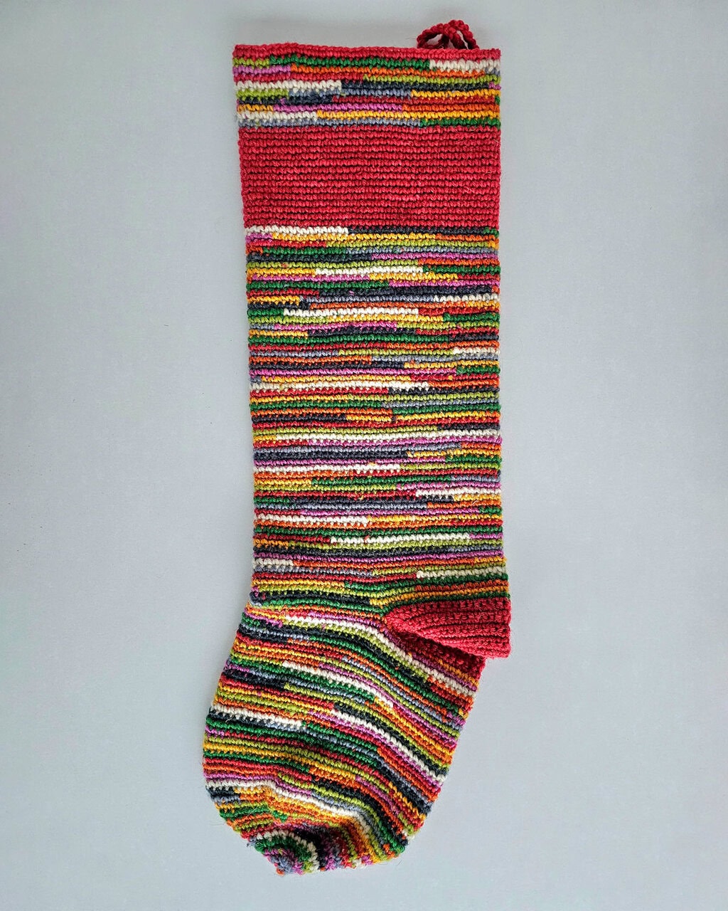 Hand-crocheted BHcrafts Christmas stocking in a vibrant rainbow stripe pattern, with a solid red cuff. Made from recycled or mixed yarns for a zero-waste design.