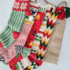 Five colorful knitted Christmas stockings with various patterns, including snowflakes, stripes, and zig-zags, arranged with a burlap stocking bag.