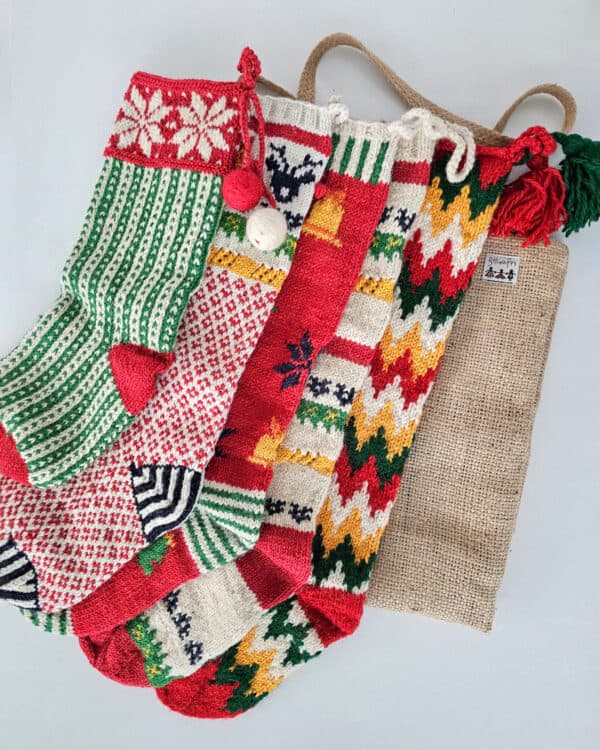 Five colorful knitted Christmas stockings with various patterns, including snowflakes, stripes, and zig-zags, arranged with a burlap stocking bag.