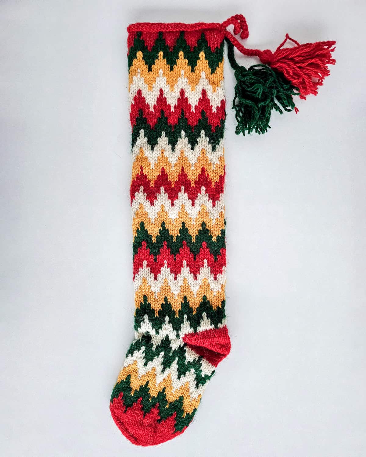 Knitted Christmas stocking with a zig-zag pattern in red, green, yellow, and white, featuring colorful tassels at the top.