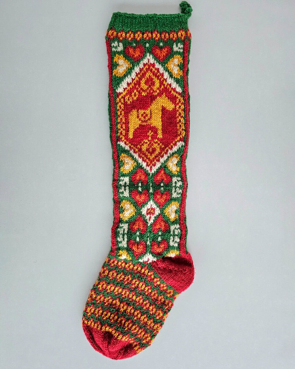 Hand-knitted BHcrafts Christmas stocking with Scandinavian-style folk motif. Features a red and green body with a prominent yellow Dala Horse design on the cuff.