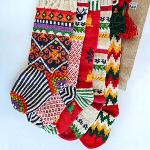 Five colorful knitted Christmas stockings with various patterns, including snowflakes, stripes, and zig-zags, arranged with a burlap stocking bag.