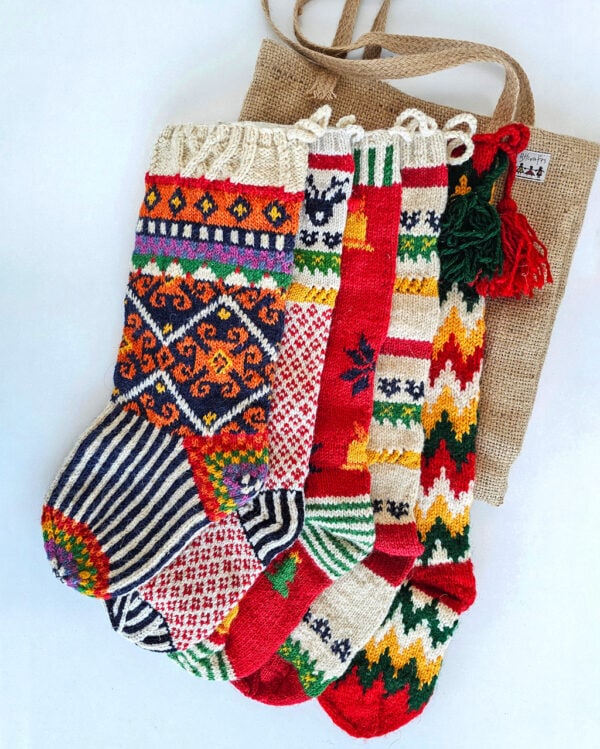 Five colorful knitted Christmas stockings with various patterns, including snowflakes, stripes, and zig-zags, arranged with a burlap stocking bag.