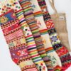 Five colourful hand-knitted Christmas stockings with folk patterns, shown with a natural jute tote bag.